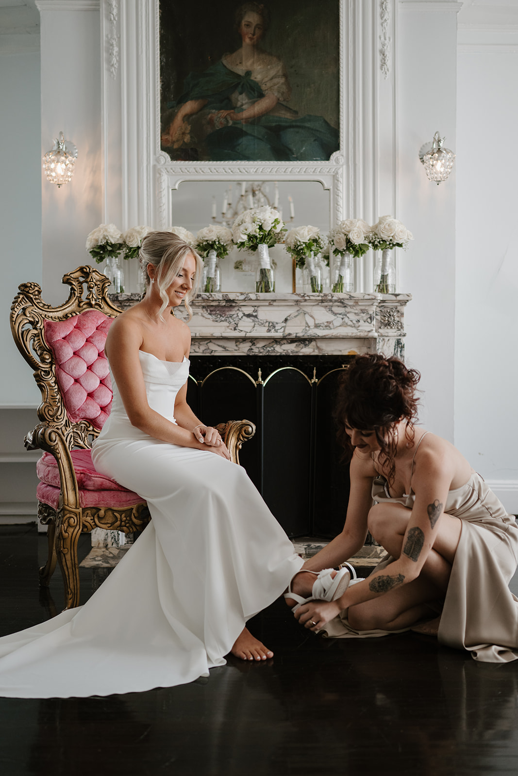 Bridesmaid helps the bride fasten her white heels as she sits on a pink velvet chair before a mantel.